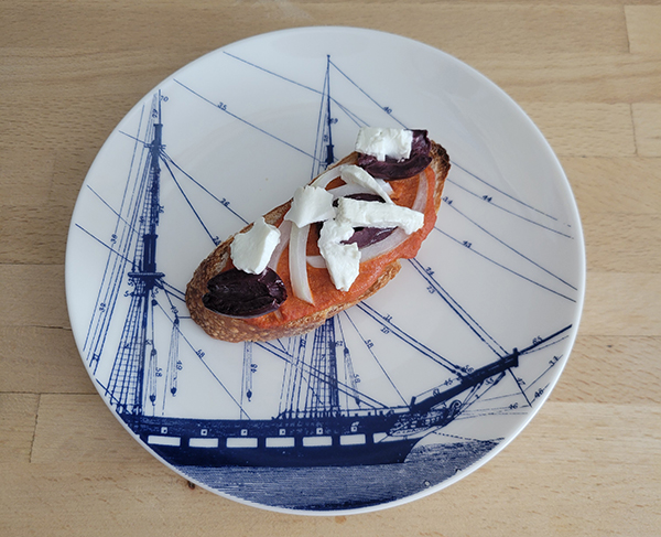 a plate with a slice of bread topped with an orange spread, feta, onions and black olives