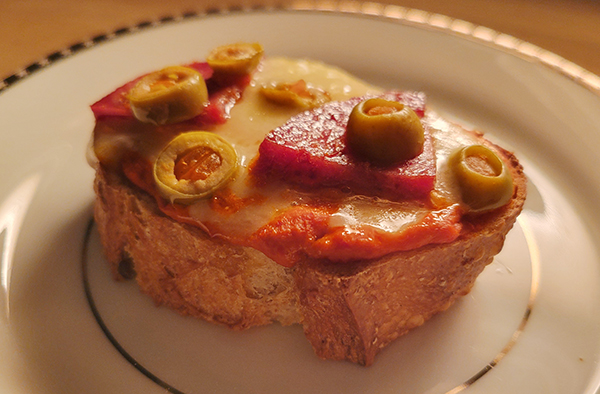 a plate with a slice of bread topped with an orange spread, salami, melted cheese and sliced green olives