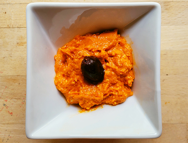 a square bowl with an orange spread and a black olive in it